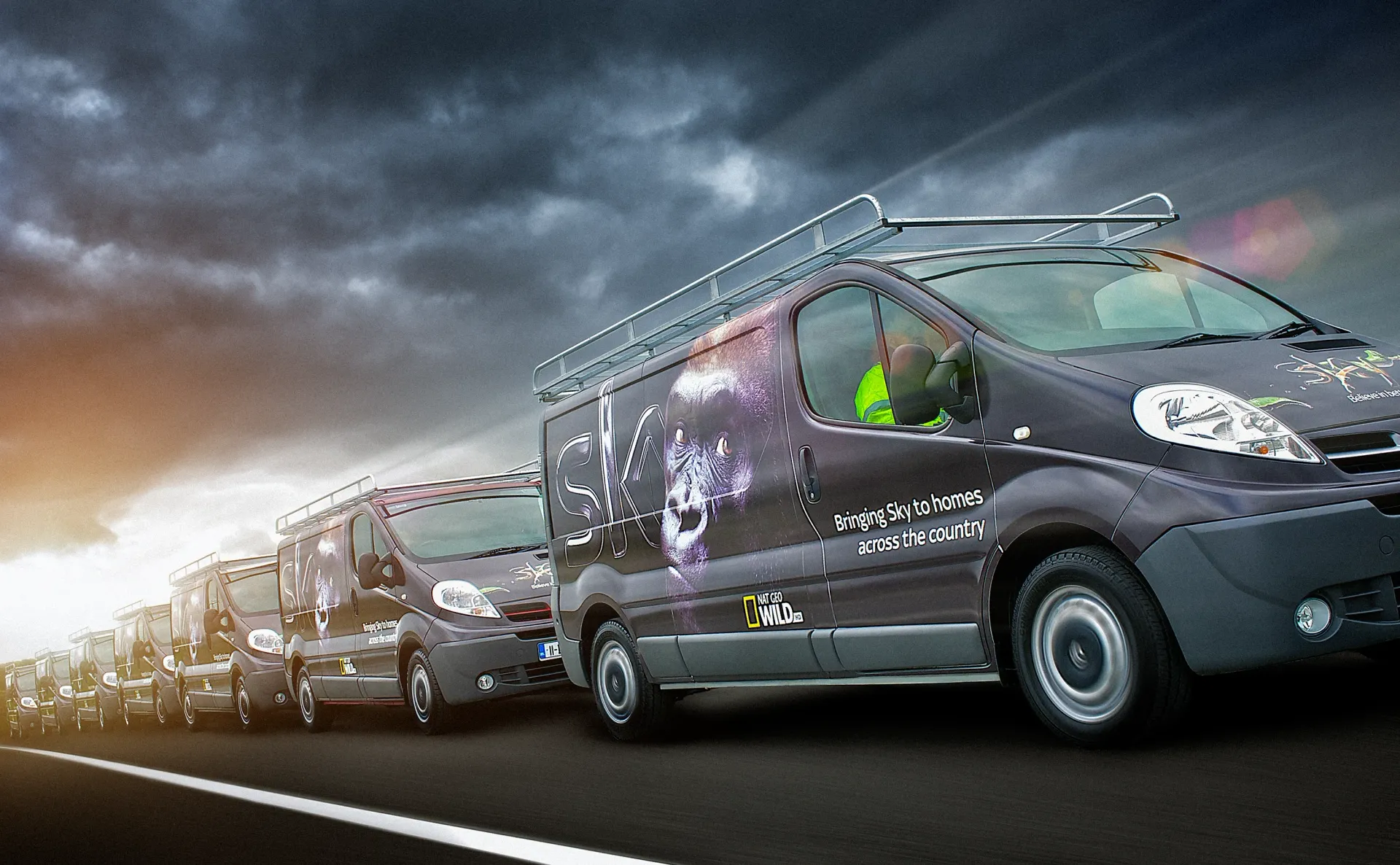 A line of dark vans with logos driving on a road under a cloudy sky.