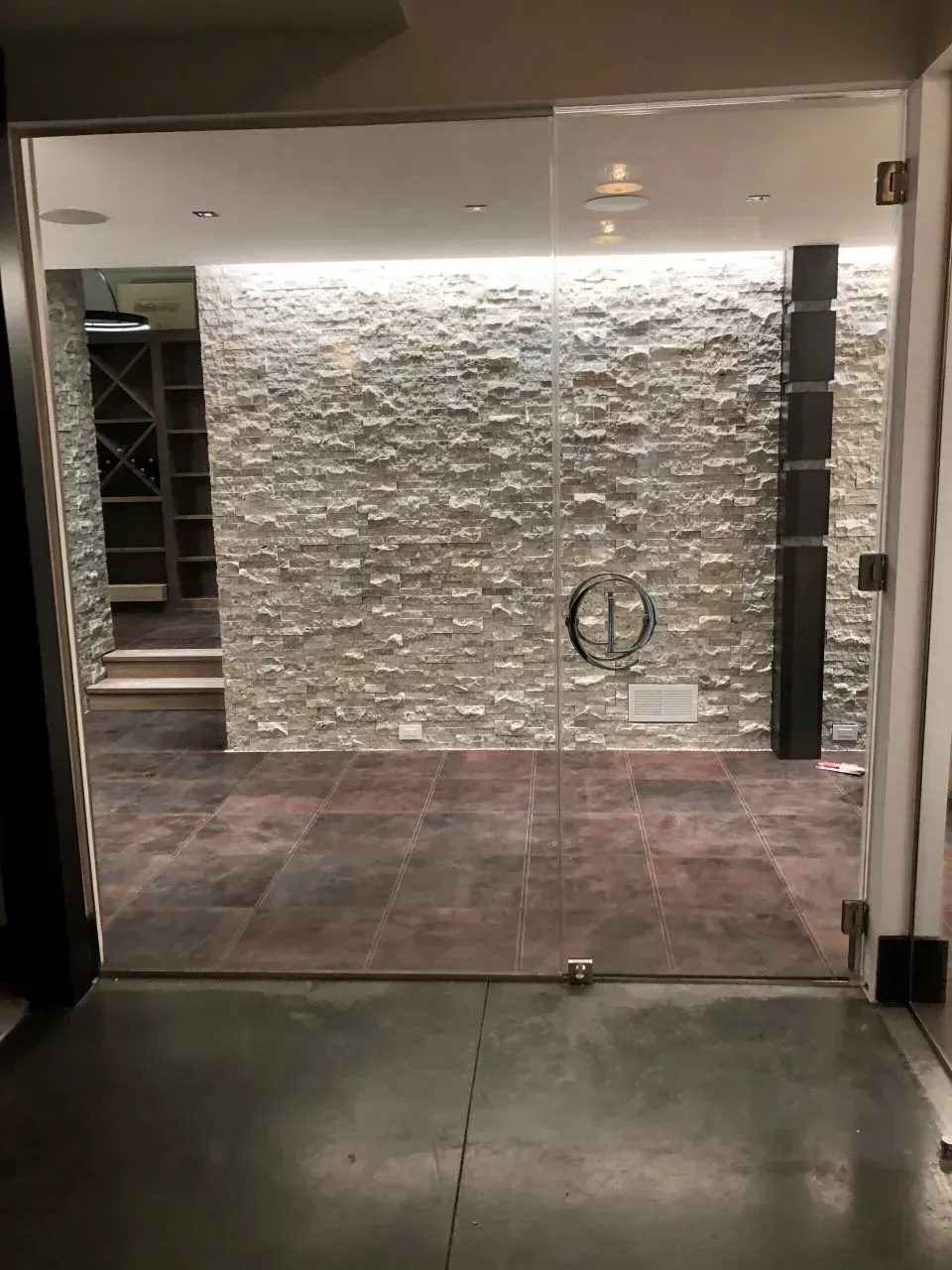 Glass door leads to wine cellar with stone wall and dark floor.