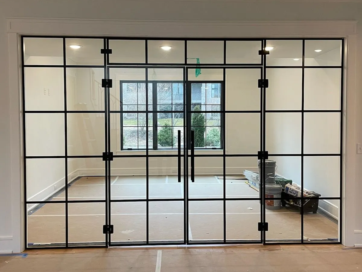 Glass-paneled doors with black frames open to a room with a window, white walls, and a paper-covered floor.