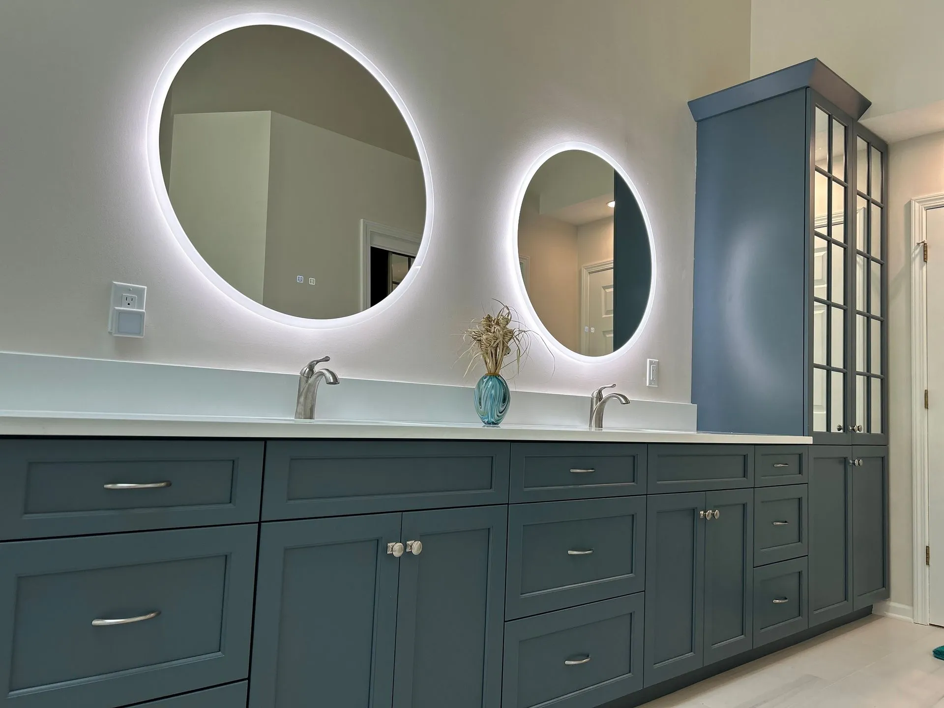 Bathroom vanity with two illuminated round mirrors, blue cabinets, white countertop, and tall cabinet.