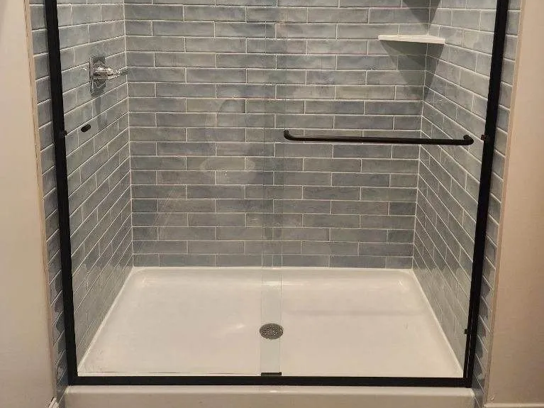 Shower with gray brick-like tile, black frame, glass door, and white base, grab bar.