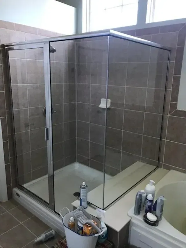 Corner shower with sliding glass doors, white base, brown tiled walls, near a tub.