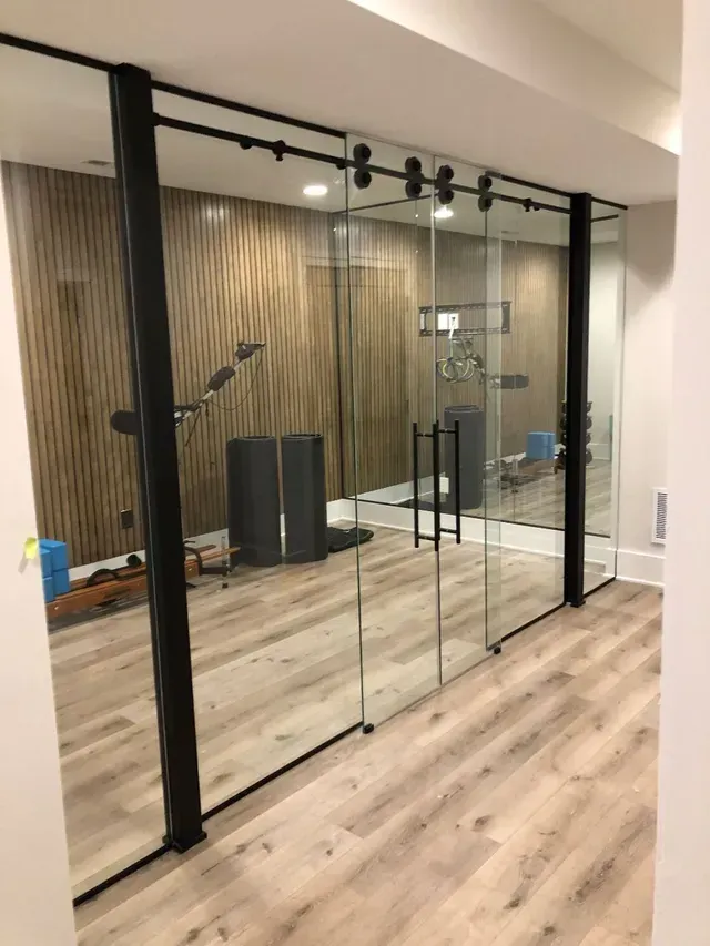 Glass sliding doors with black framing in a home gym. Wooden wall paneling and exercise equipment visible inside.