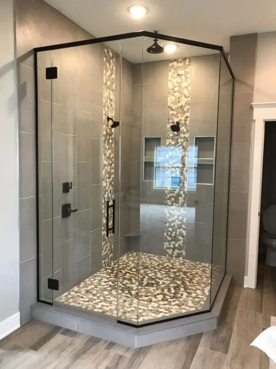 A modern glass corner shower with black framing, gray tile, and pebble flooring.