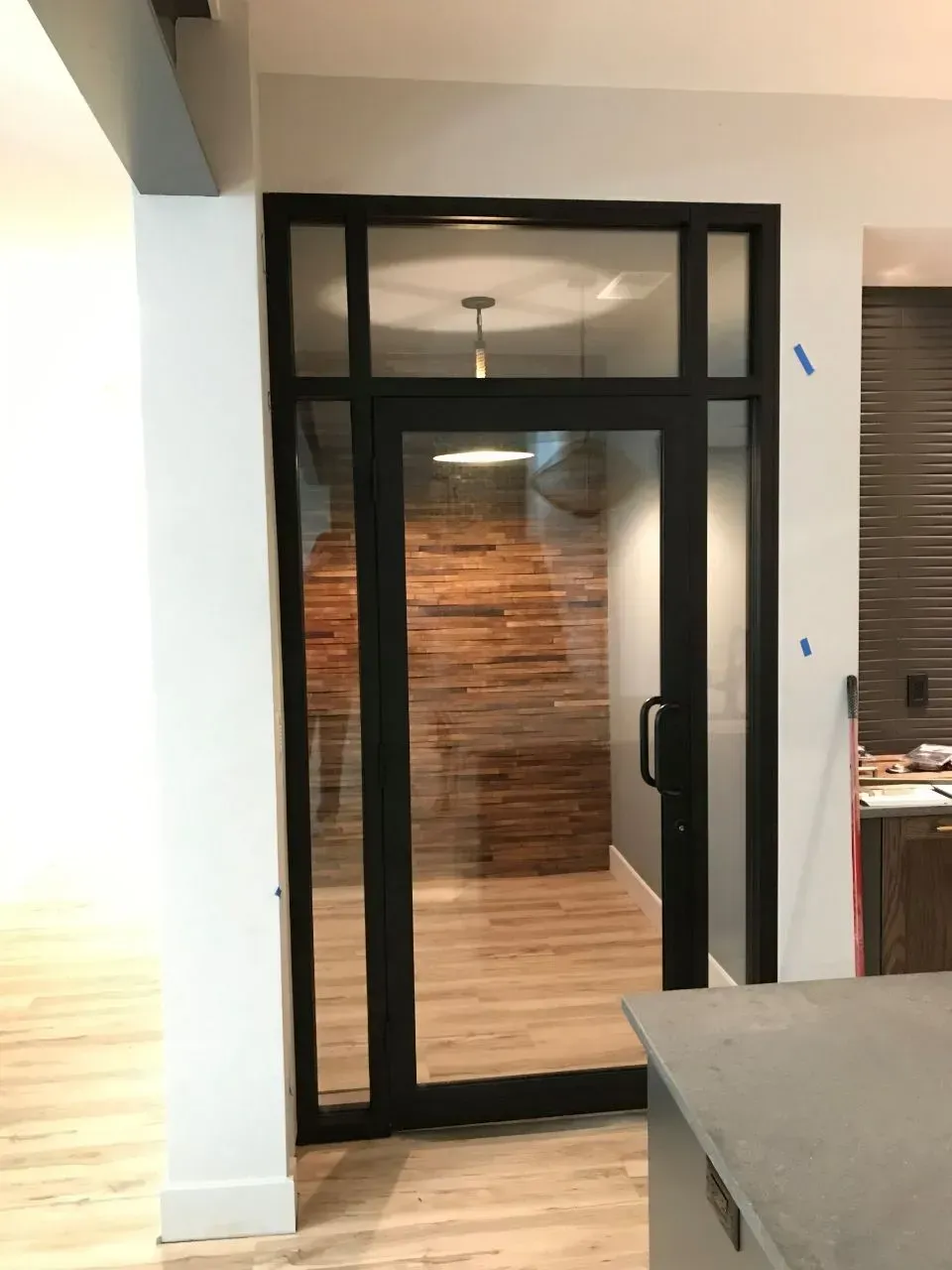 Black framed glass door with a transom, leading to a room with a wooden accent wall.