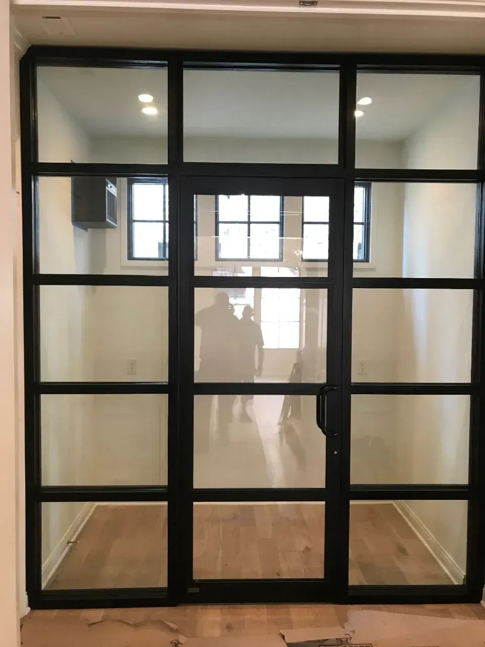 Black-framed glass door with multiple panes, revealing a room with people inside. Wood floor, white walls.