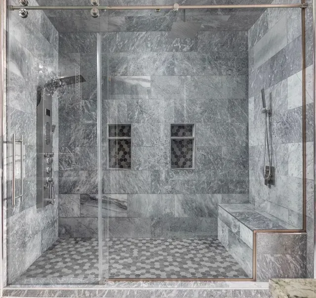Gray tiled shower with glass door, pebble floor, built-in bench, and two recessed shelves.