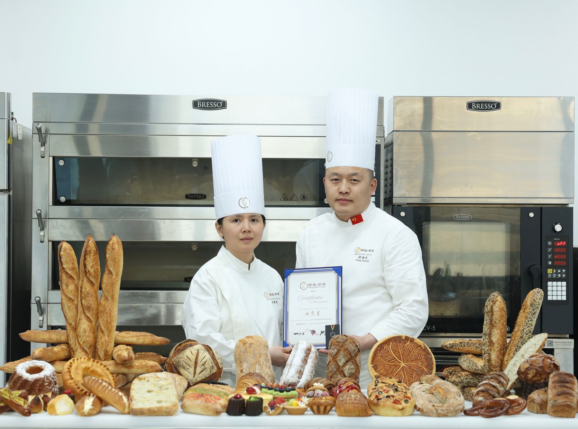 Two chefs in white uniforms with various bread types, holding a certificate in a kitchen setting.