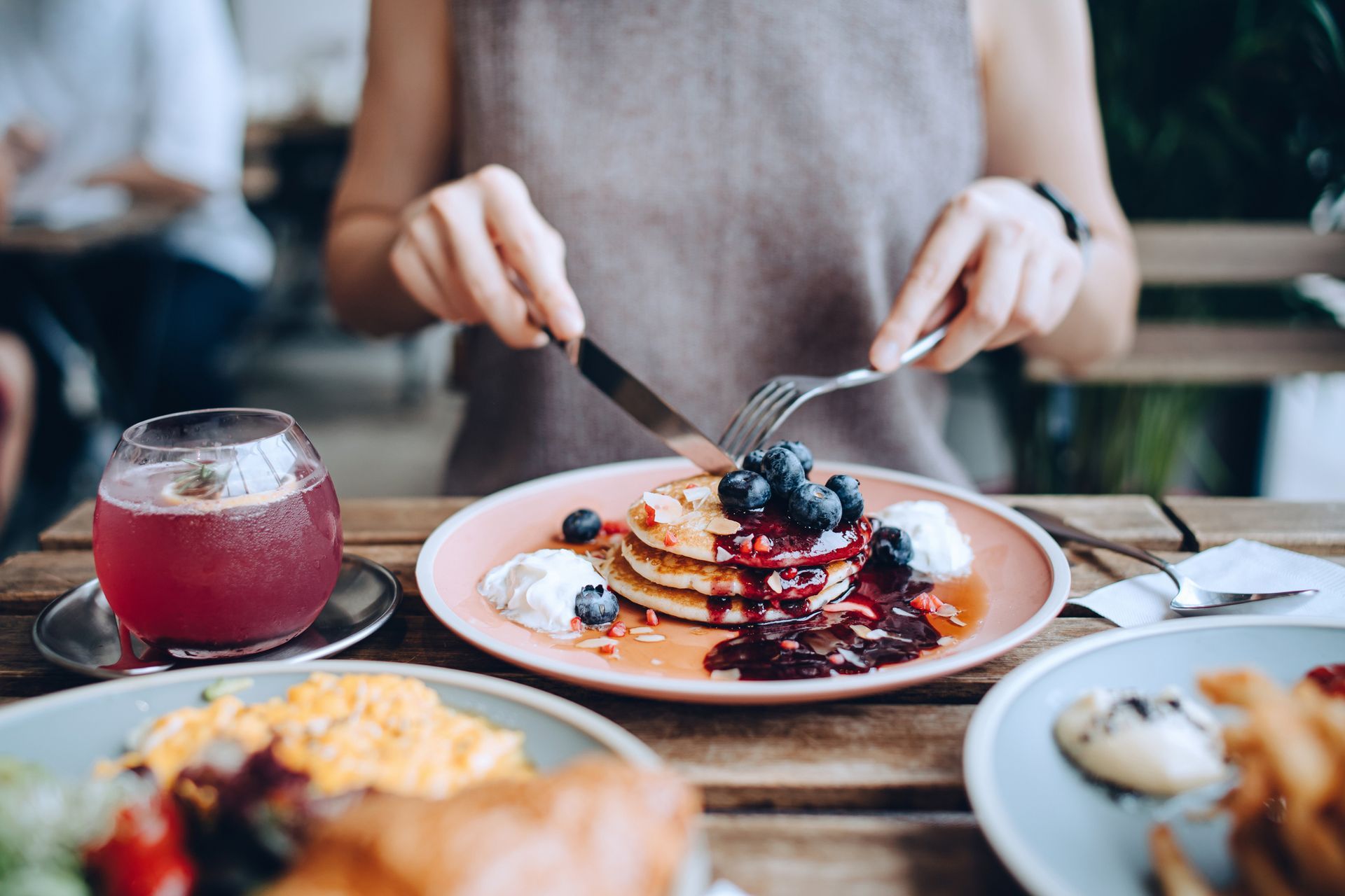 Close up of young woman sitting at dining table eating pancakes with blueberries and whipped cream