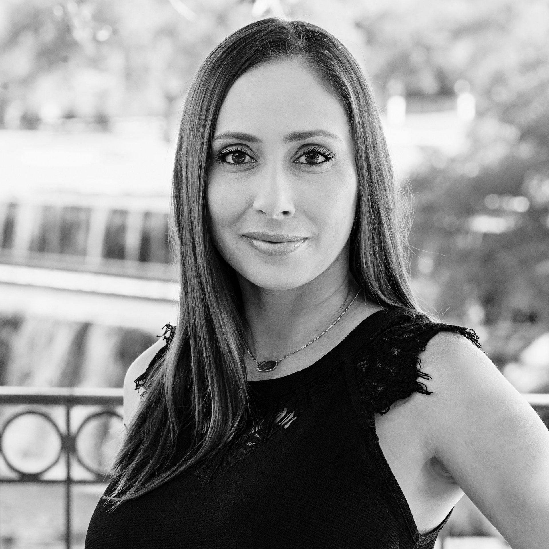 Woman with long hair in black dress, smiling outdoors near a building with water features.