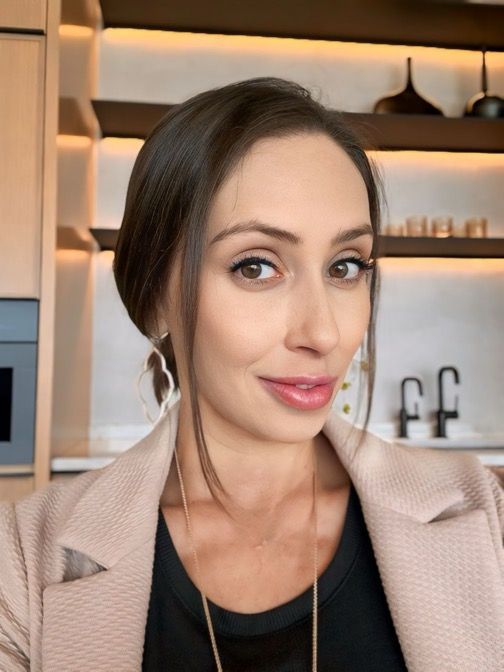 Woman in blazer, dark top, with earrings, smiling. Kitchen with shelves in background.
