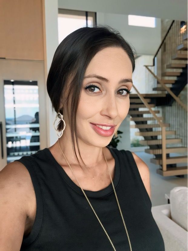 Woman with dark hair, black tank top, smiling in a modern home with stairs and outdoor view.