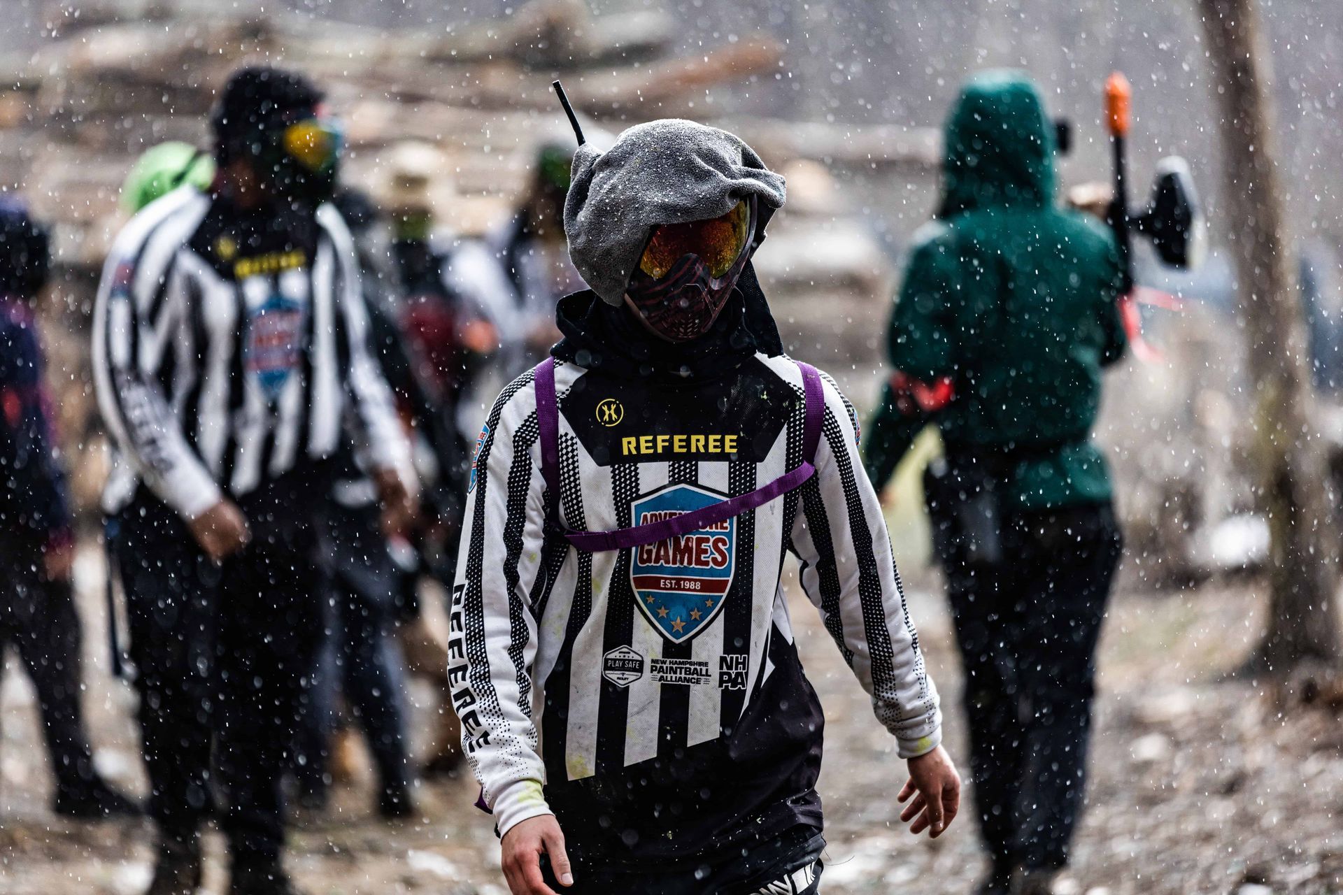 People in paintball gear outdoors in a rainy setting, one wearing a striped referee jersey.