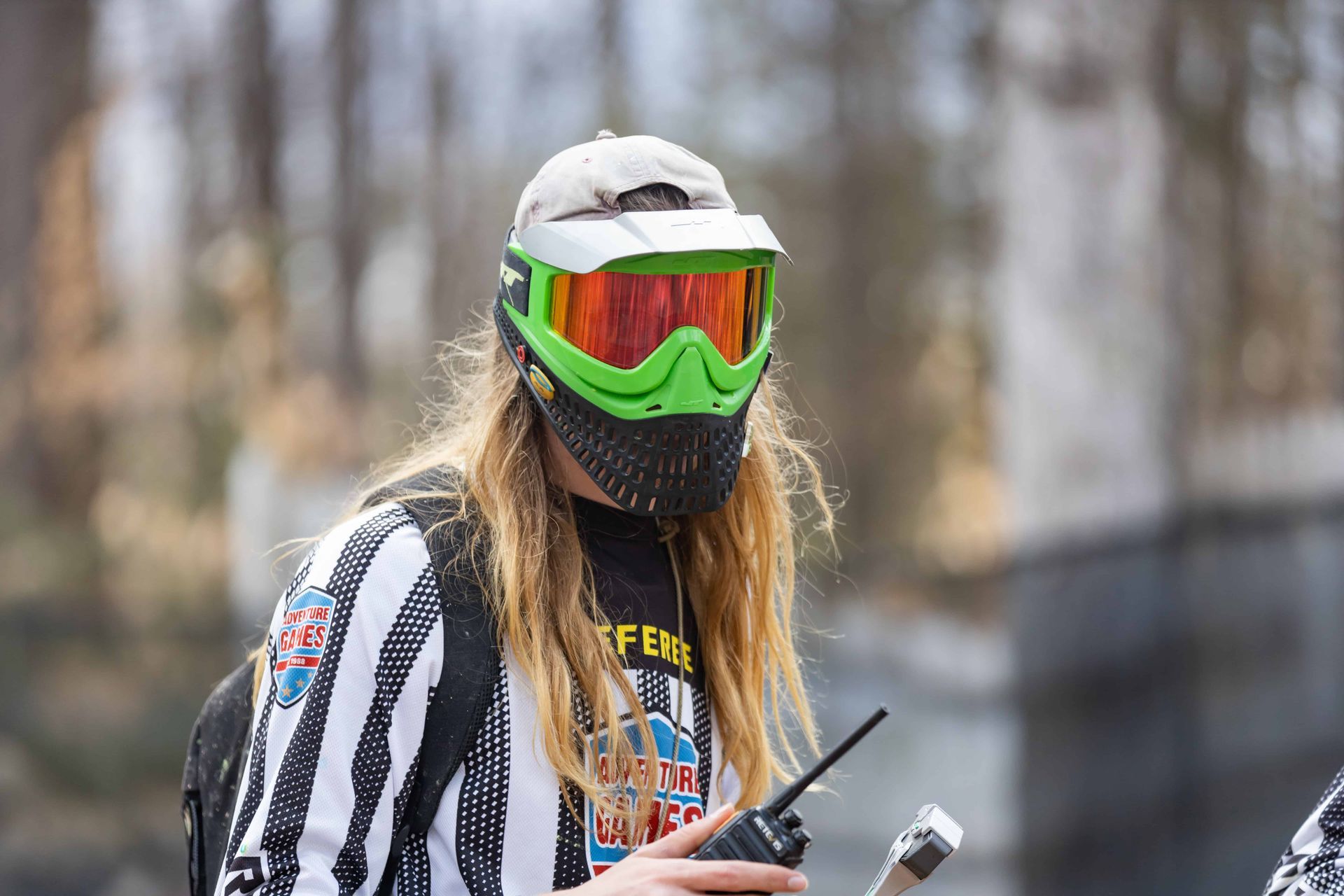 Person in paintball mask holding a radio. Wearing a striped jersey and baseball cap, outdoors.