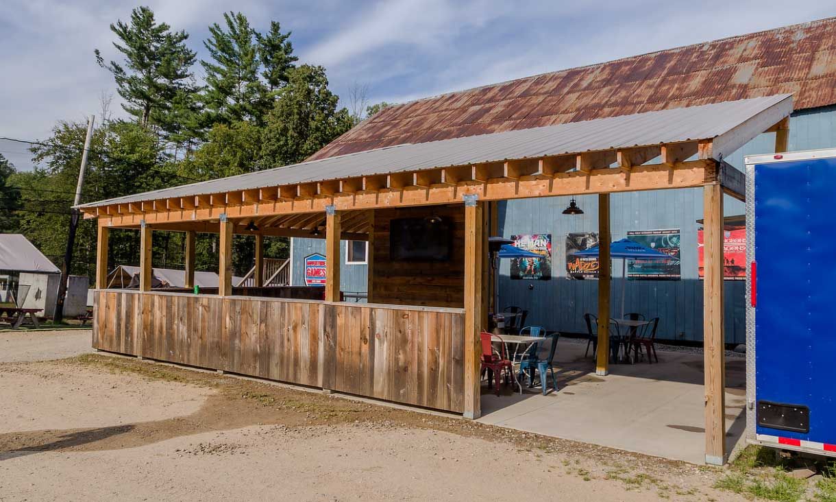 Outdoor bar with wooden facade and corrugated metal roof. Tables, chairs, and signs visible.