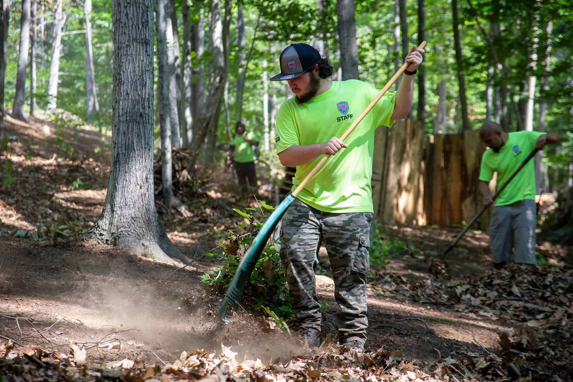Men raking leaves in a forest. They wear bright green shirts and hats. Sunshine streams through the trees.