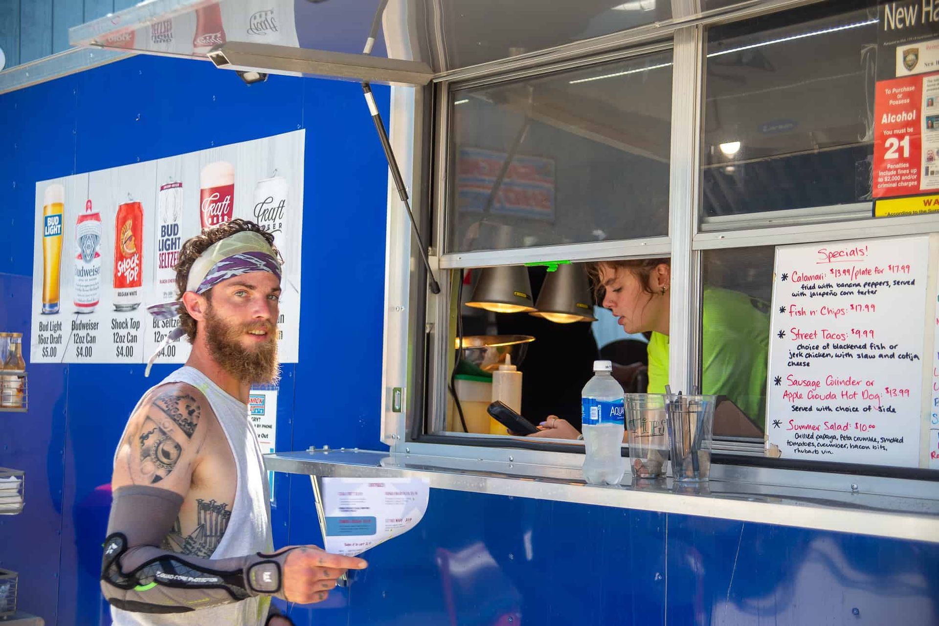 Man ordering from food truck; he's holding a menu, looking at the camera. Another person inside the truck takes the order.