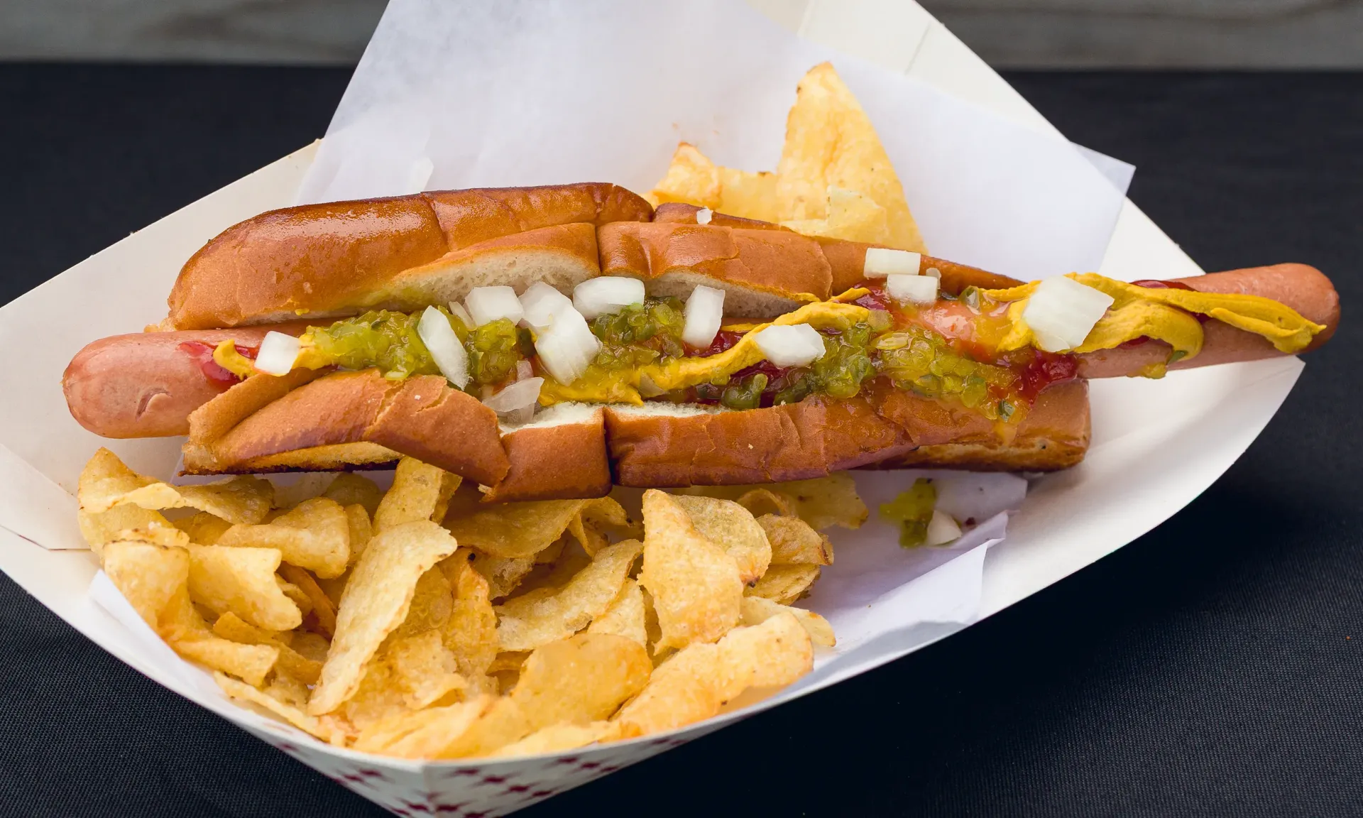 Hot dog in a split bun with toppings, served with potato chips in a paper tray.