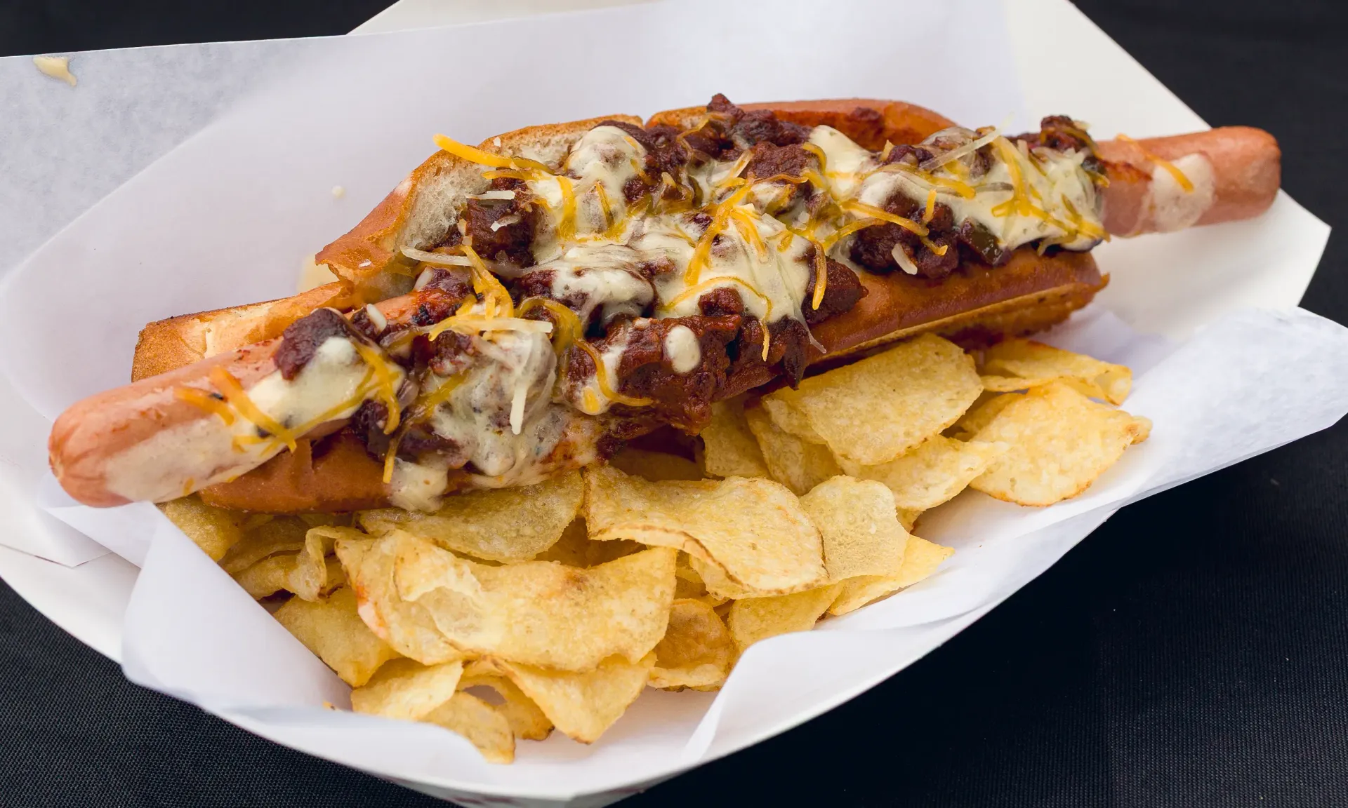 Chili cheese hot dog on a toasted bun with chips in a white paper tray.