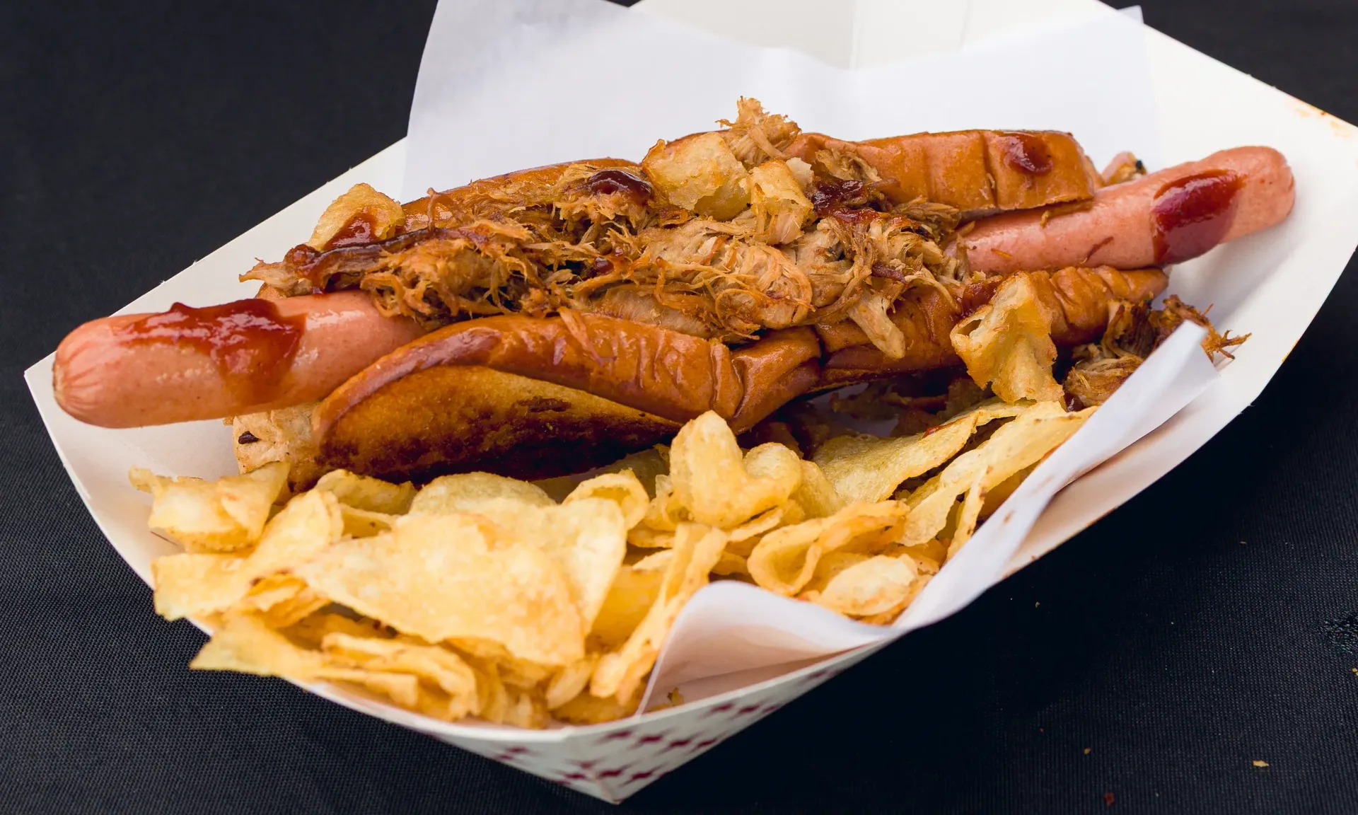 Hot dog with pulled pork, chips, and barbecue sauce in a paper tray.