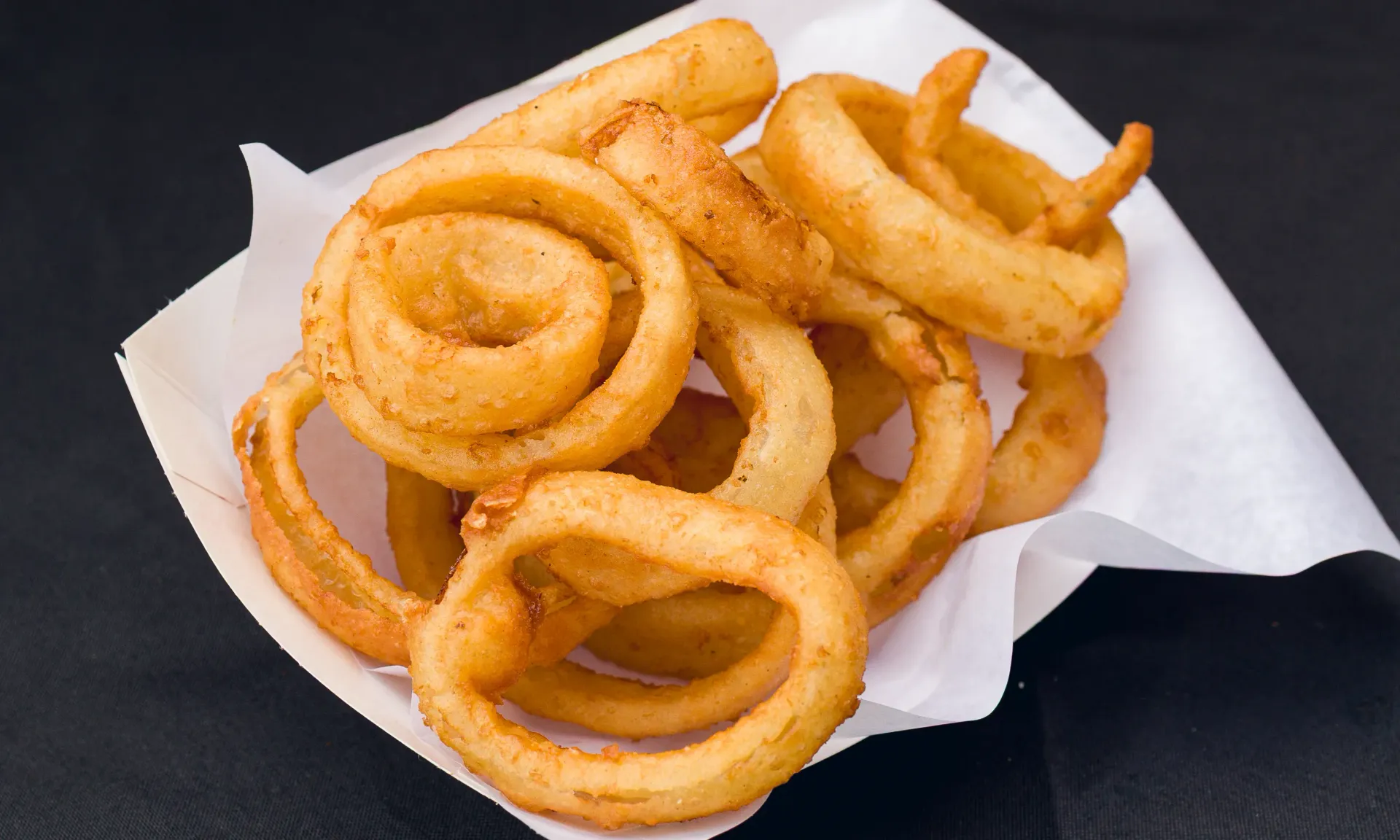 Curly fries in a white paper container on a black surface.