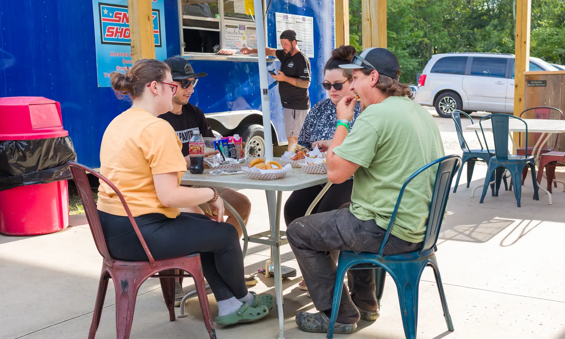 Four people sit at an outdoor table, eating and talking. A food truck is visible in the background.