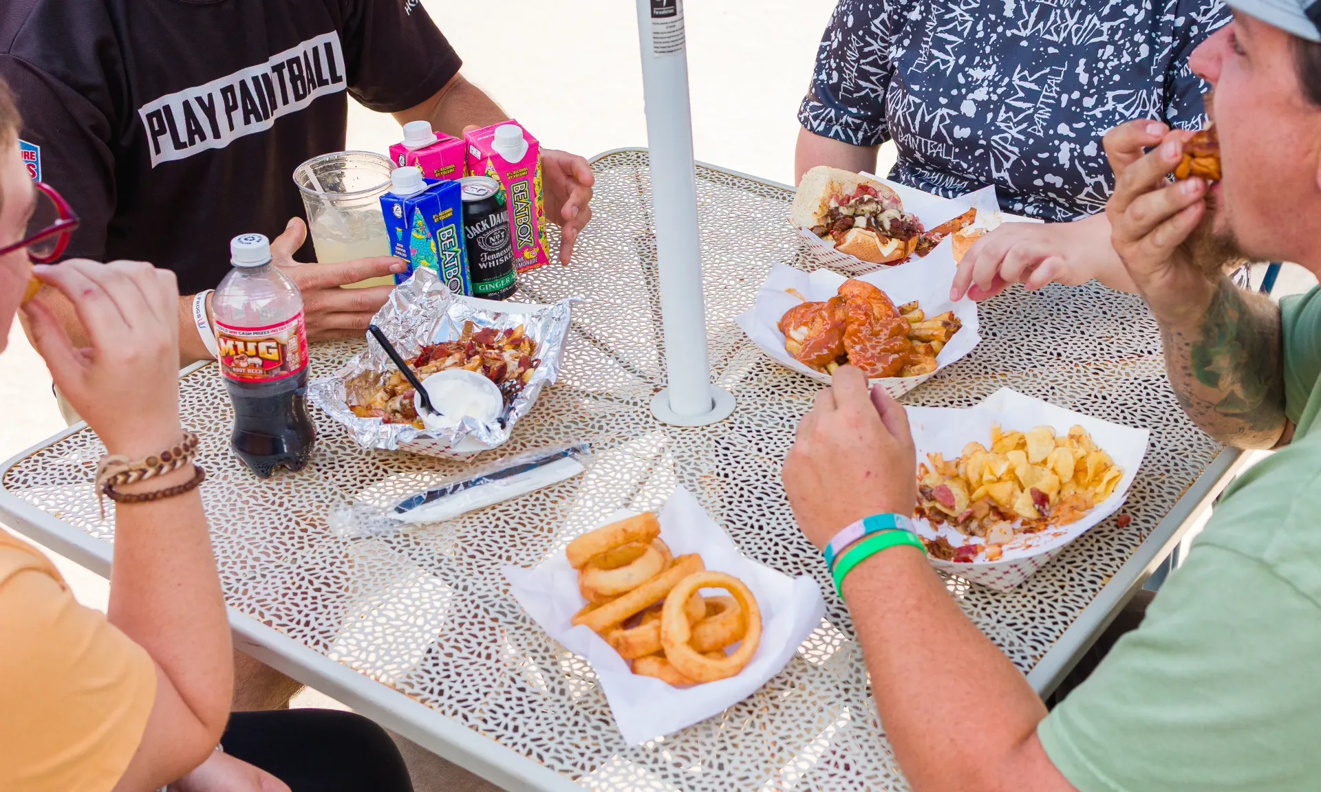 People eating at an outdoor table with various food items like onion rings, wings, and mac and cheese.