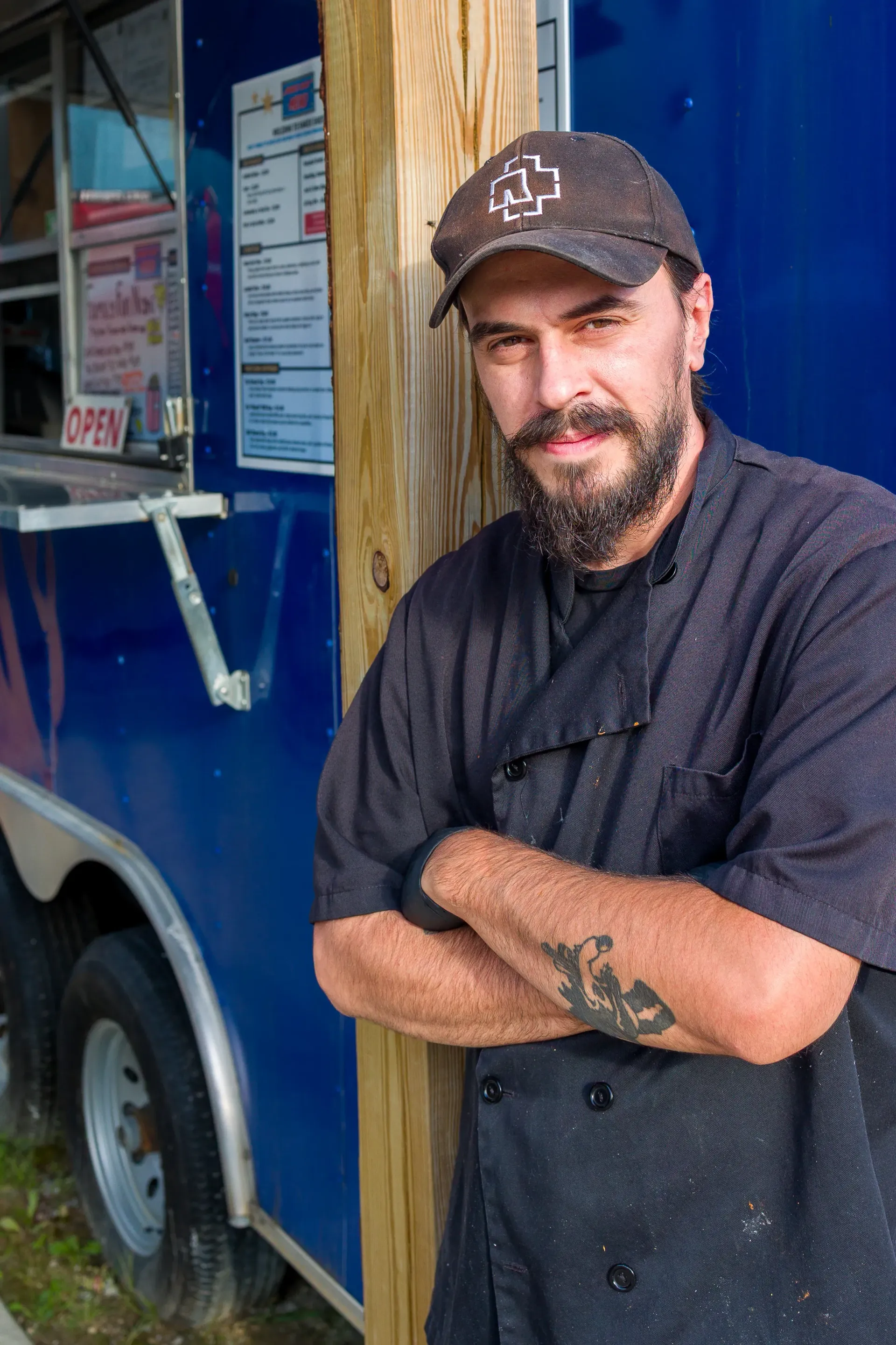 Man with beard and chef's uniform leans against a food truck; smiling, arms crossed.