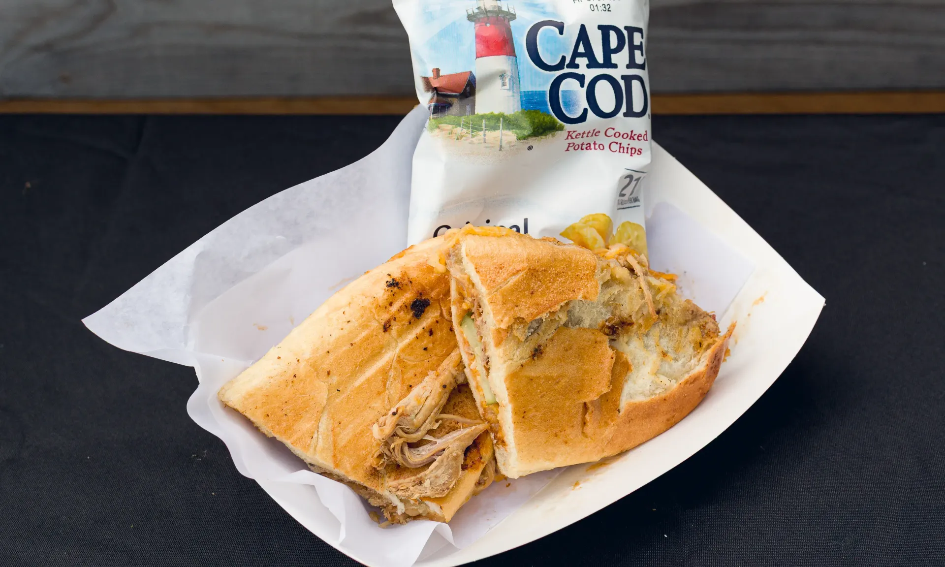 Cuban sandwich on a paper plate with a bag of Cape Cod chips, on a dark table.