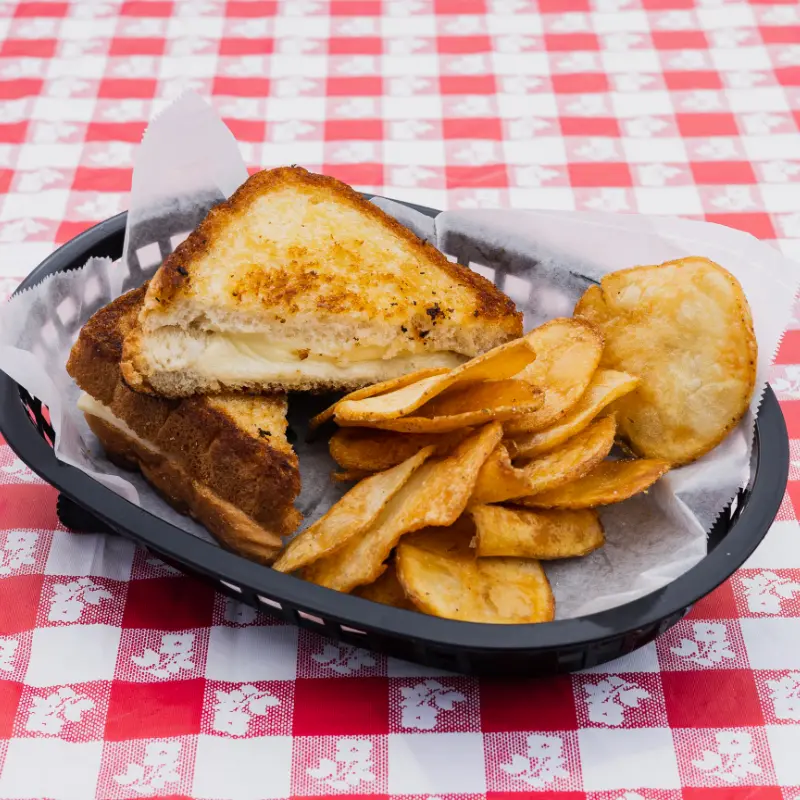 Grilled cheese sandwich with potato chips in a black basket on a red and white checkered tablecloth.