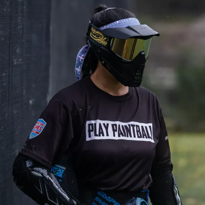 Person in paintball gear, wearing a mask and visor, standing near netting.