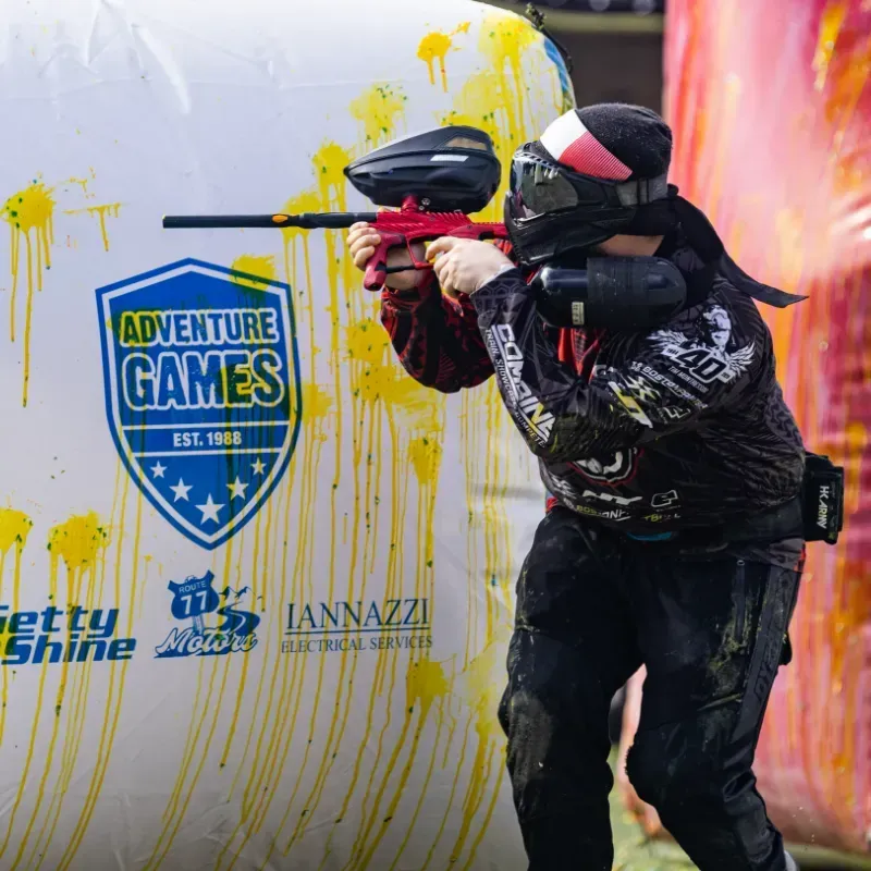 A paintball player aims a red gun from behind a bunker.  They wear black gear and a face mask, at Adventure Games.