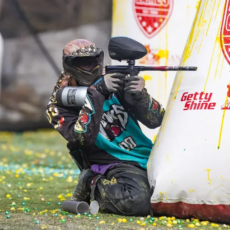 Paintball player aiming gun behind a bunker on a green field.