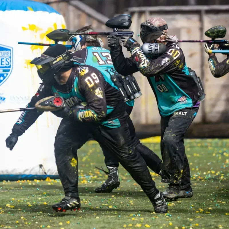 Paintball players in turquoise and black gear aiming guns on a green field.