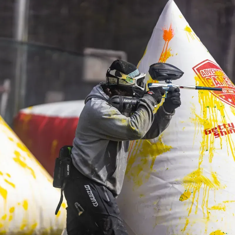 A paintball player aiming his marker behind a white bunker, wearing protective gear and holding a marker.