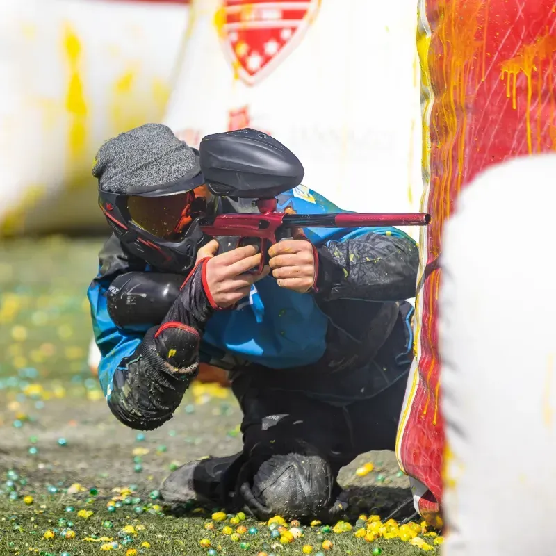Paintball player crouched behind a barrier, aiming a red paintball gun. Yellow and blue paint splatters.