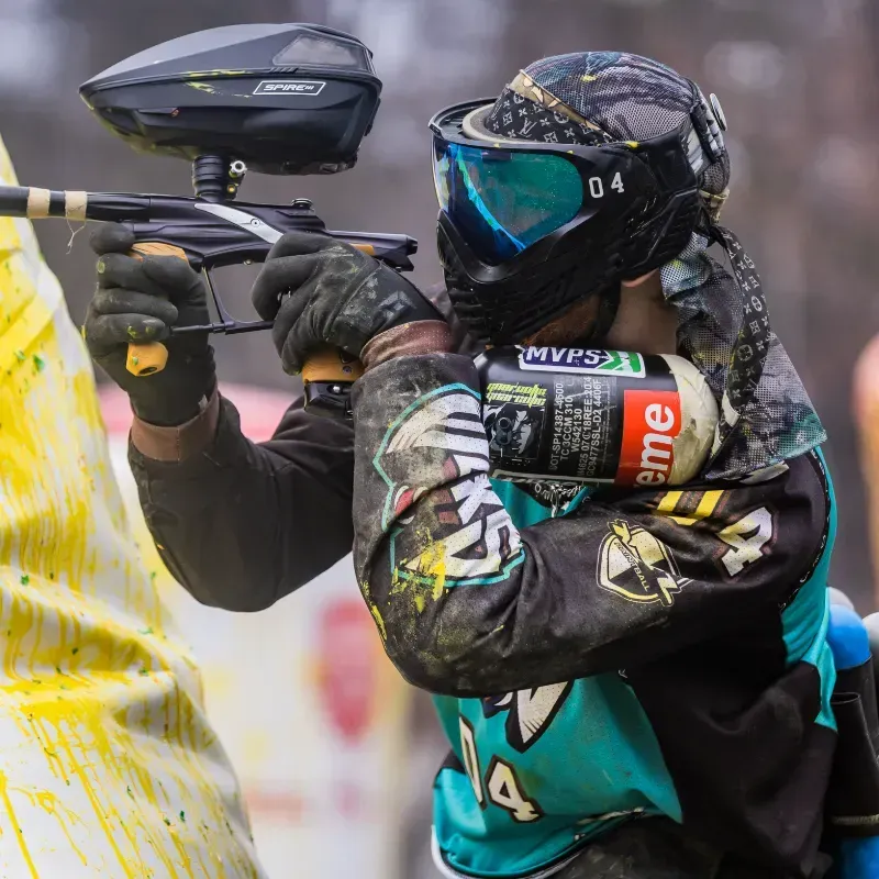 Paintball player aiming marker, wearing blue and black gear, in outdoor game setting.