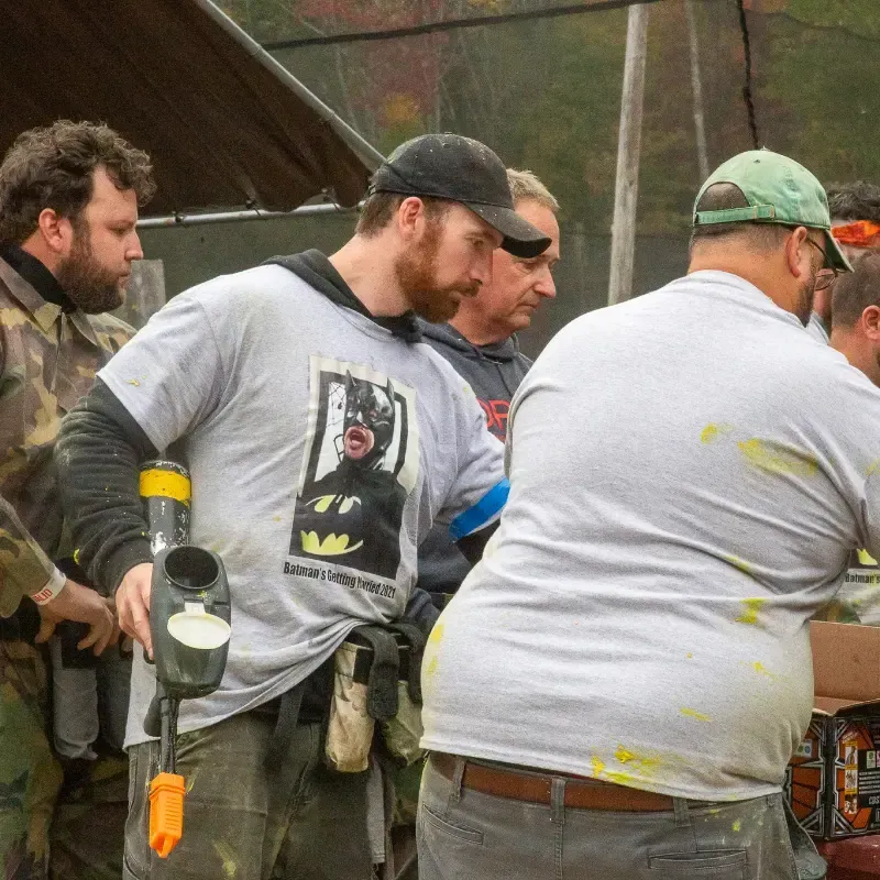 Men in paintball gear preparing equipment outdoors. One wears a Batman shirt.