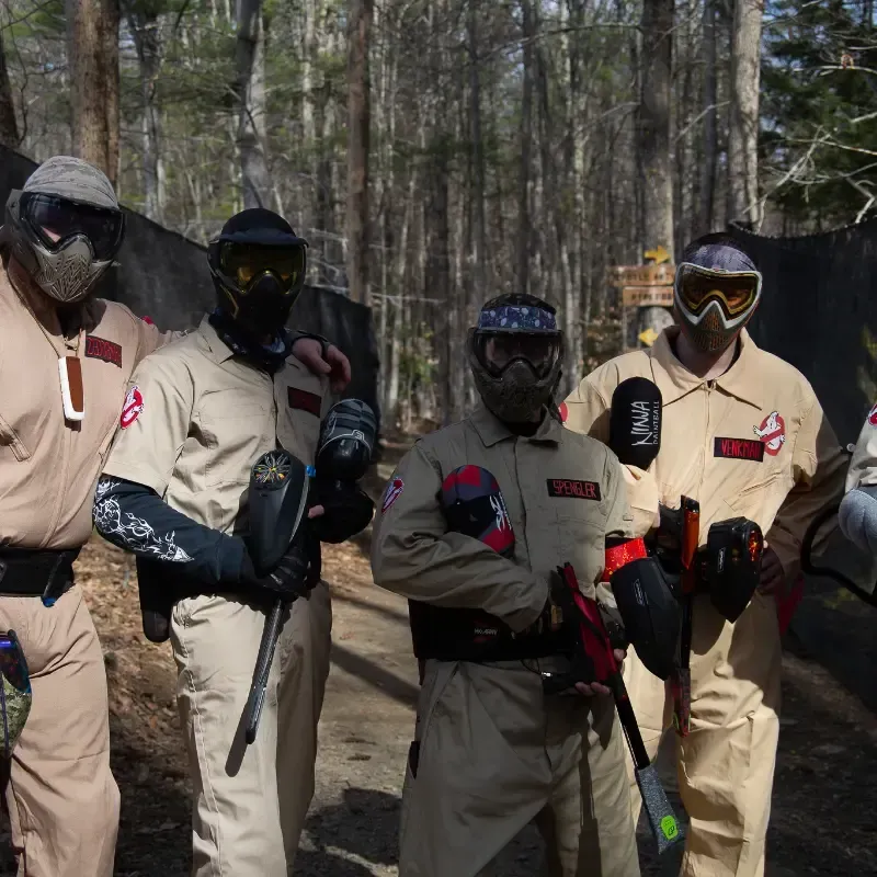 Four people in Ghostbusters costumes holding paintball guns in a wooded area.