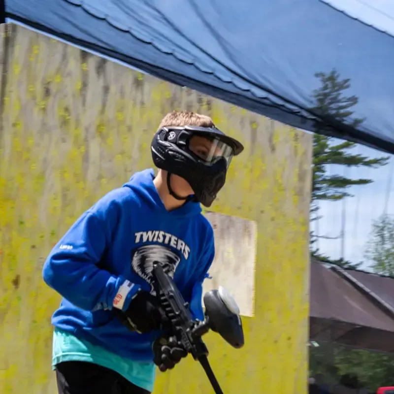 Boy in blue hoodie and paintball mask aiming a paintball gun outdoors.