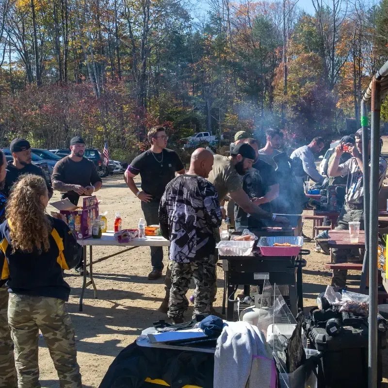 A group of people grilling food outside, surrounded by tables and cars on a sunny day.