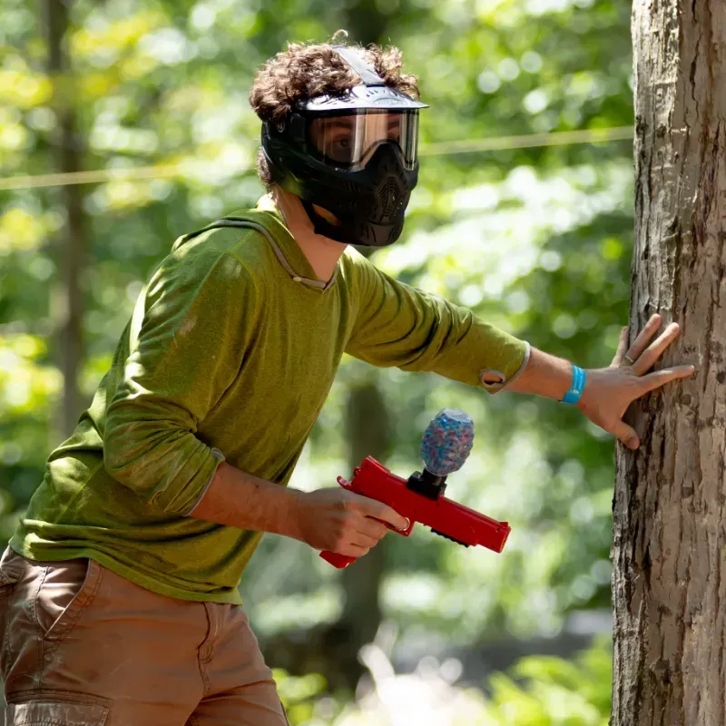 Man in paintball mask and green shirt, aiming a water-based gel blaster by a tree in a wooded area.