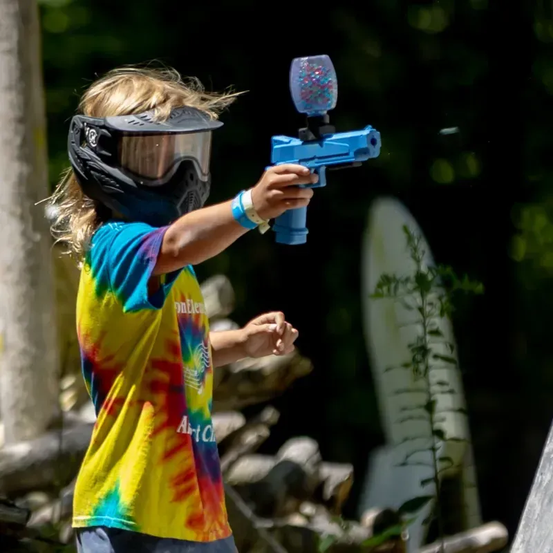 Child in tie-dye shirt and paintball mask aims a blue paintball gun outdoors.