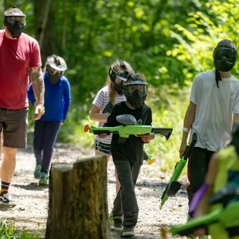 People in paintball masks walk through a wooded area, holding paintball guns.