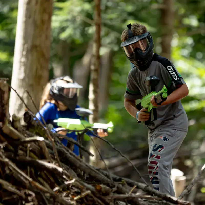 Two people in paintball gear aiming guns in a wooded area, behind a pile of logs.