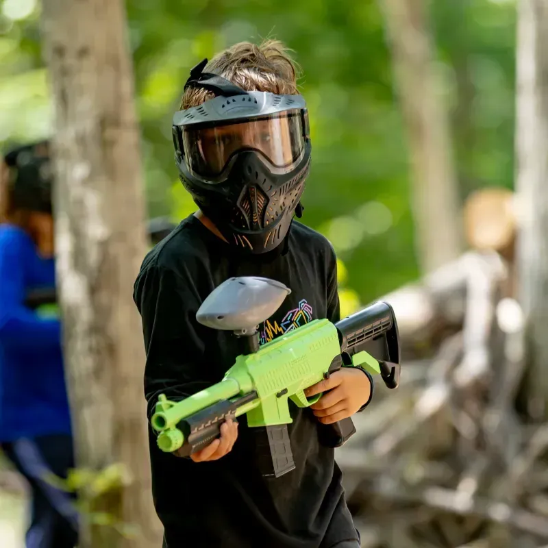 Boy in paintball mask holding a neon green paintball gun, standing outdoors in a wooded area.
