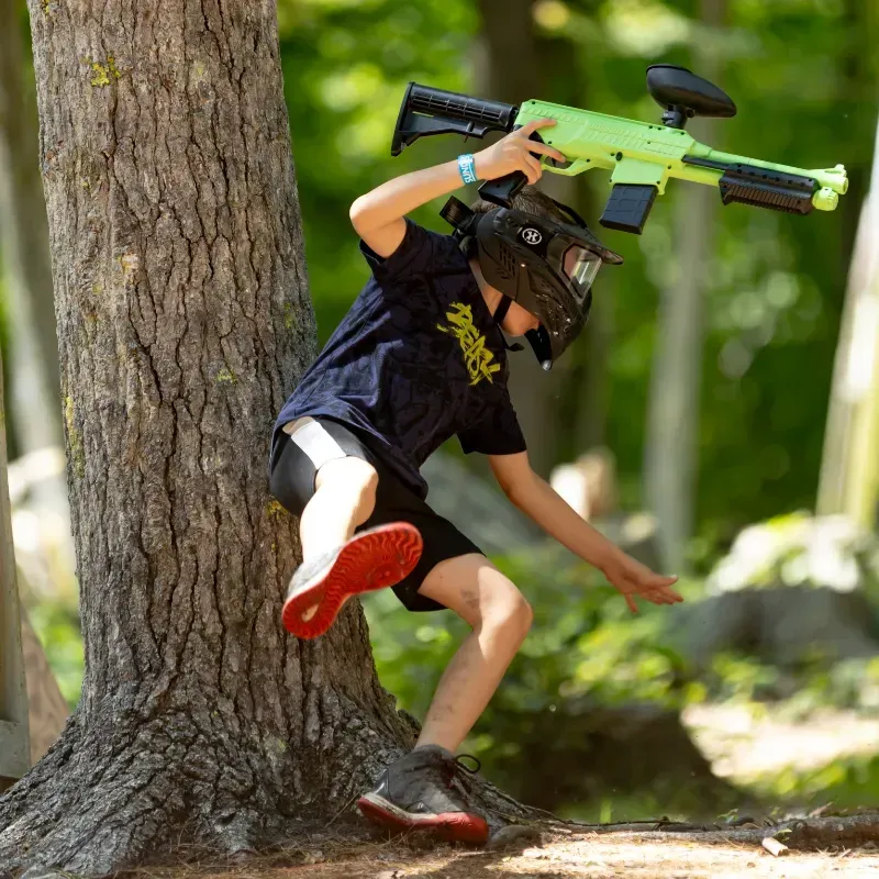 Person in paintball gear jumps near a tree, holding a neon green paintball gun in a wooded area.