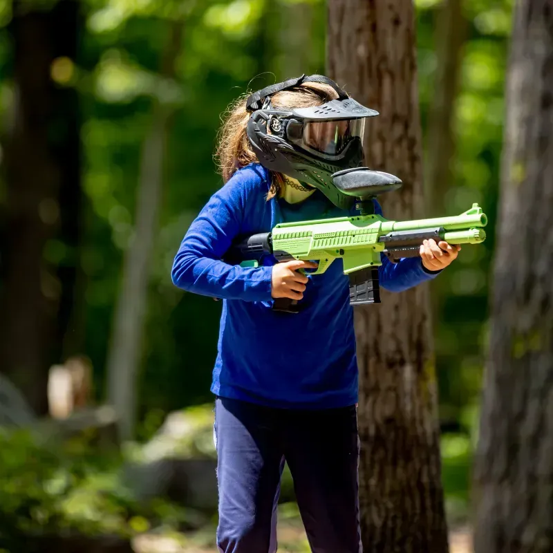 Young person with a helmet and a green paintball gun in a wooded area.