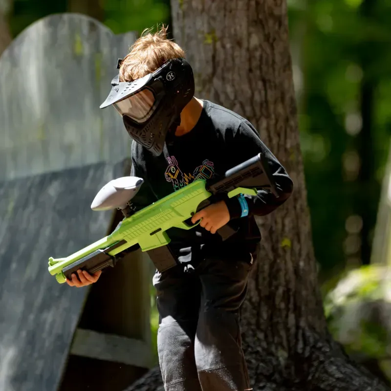 Boy wearing paintball gear aiming a bright green gun in a wooded area.