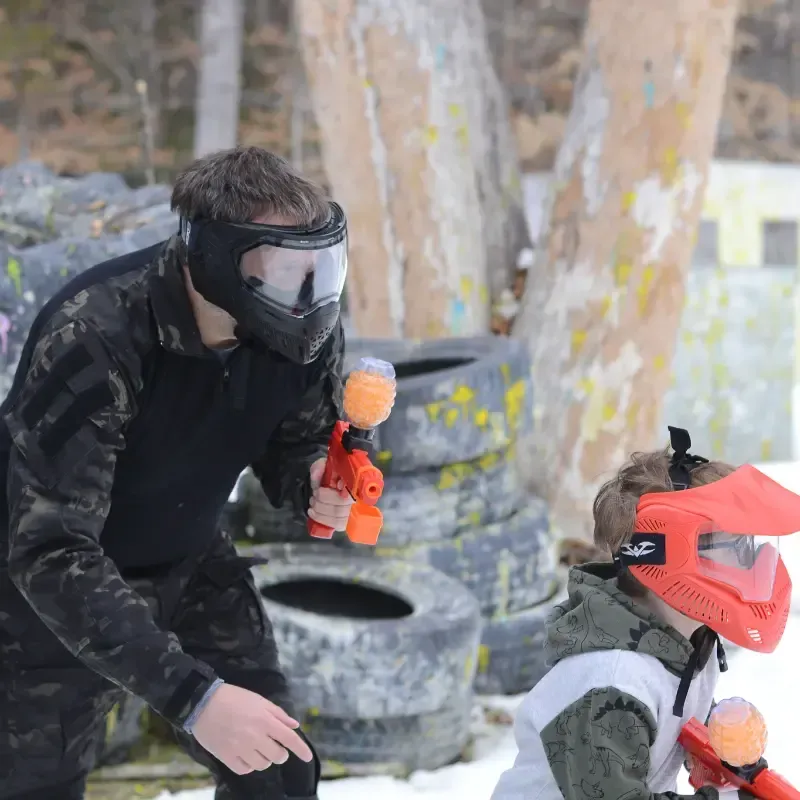 Man and child play paintball in a snowy outdoor area. Man wears a black camo suit,